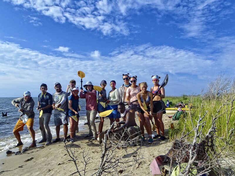 A group of people stand on a sandy beach, holding paddles and kayaks. They appear to be preparing for or returning from a kayaking trip. The sky is blue with scattered clouds, and there's vegetation on the right side of the frame. The overall scene suggests an outdoor recreational activity.